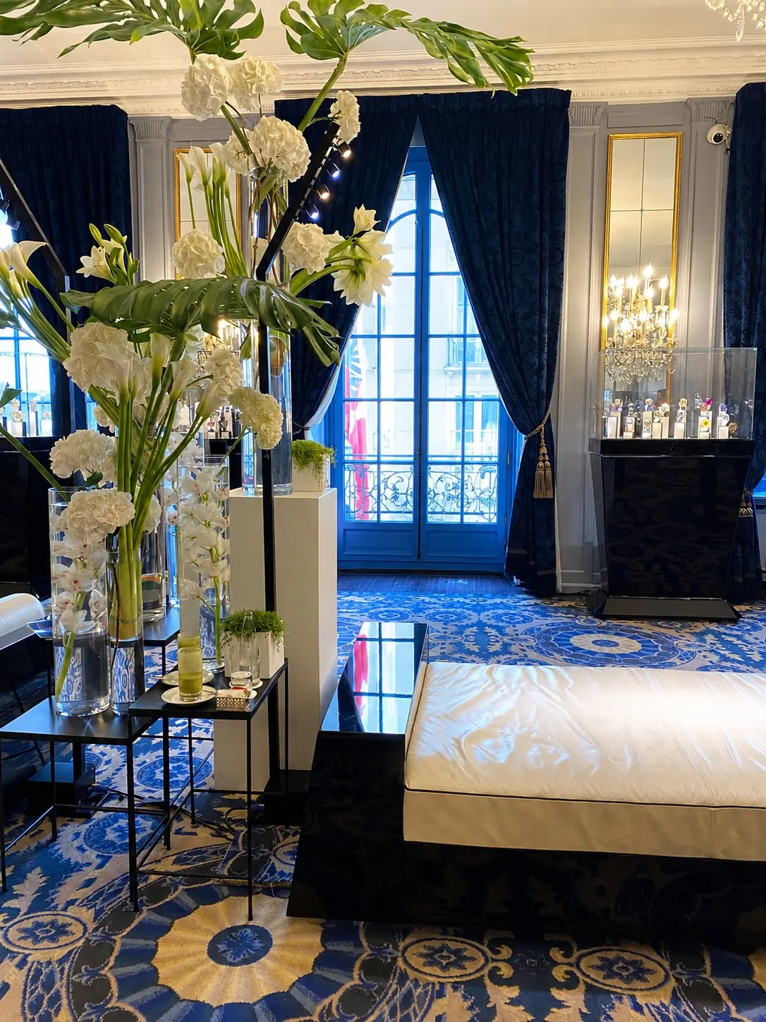 A private presentation salon: watches on black plinths, blue drapery and white flowers at the windows.