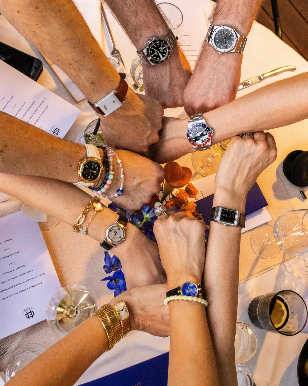 Guests around a dinner table with their wrists together, each wearing a different watch.